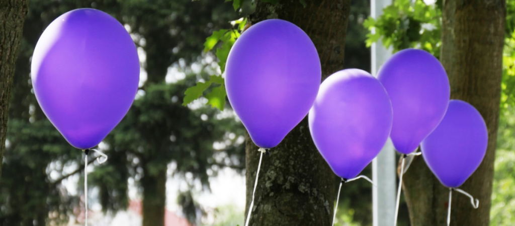 5 tethered purple balloons floating in front of a row of trees