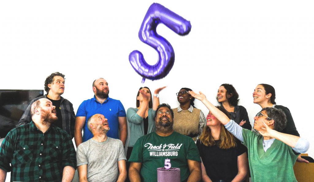 Seated in a team photo formation, the Matter of Focus team taking great pleasure in playing with a large foil 5 balloon.