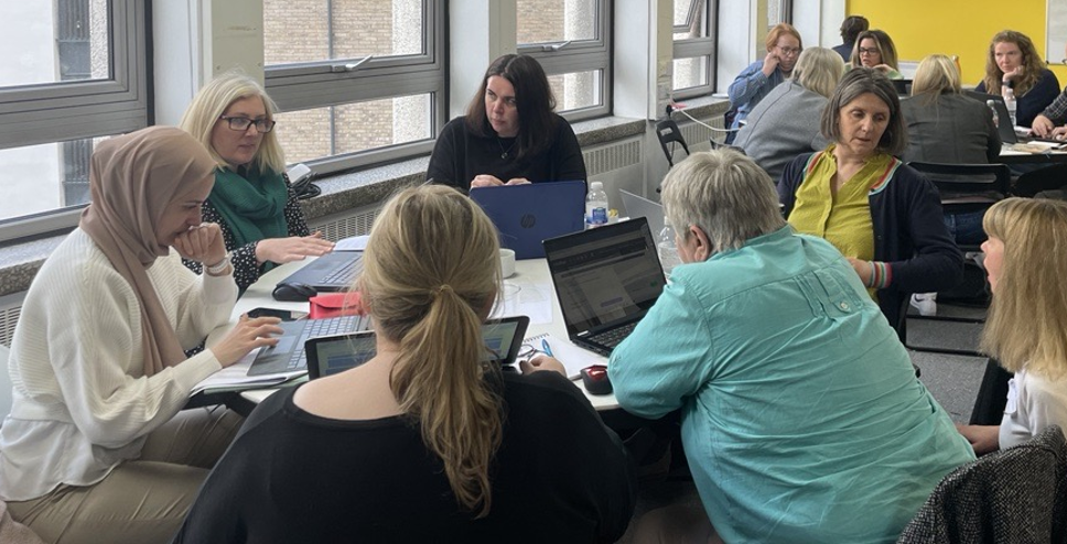 Participants at a workshop sit in small groups around tables, working on laptops and engaging in discussion. The room is bright with large windows, and the atmosphere appears collaborative and focused.